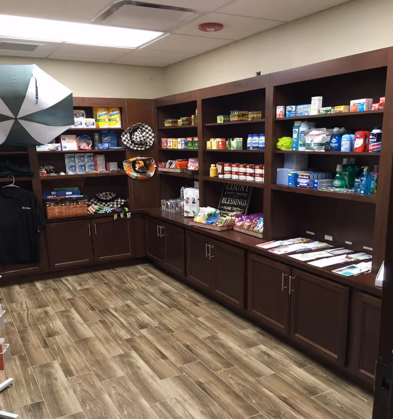Interior view of a small retail or convenience area with dark wooden shelves and cabinets stocked with various items including snacks, personal care products, hats, and clothing. The floor has wood-like tiles and the ceiling has recessed lighting.