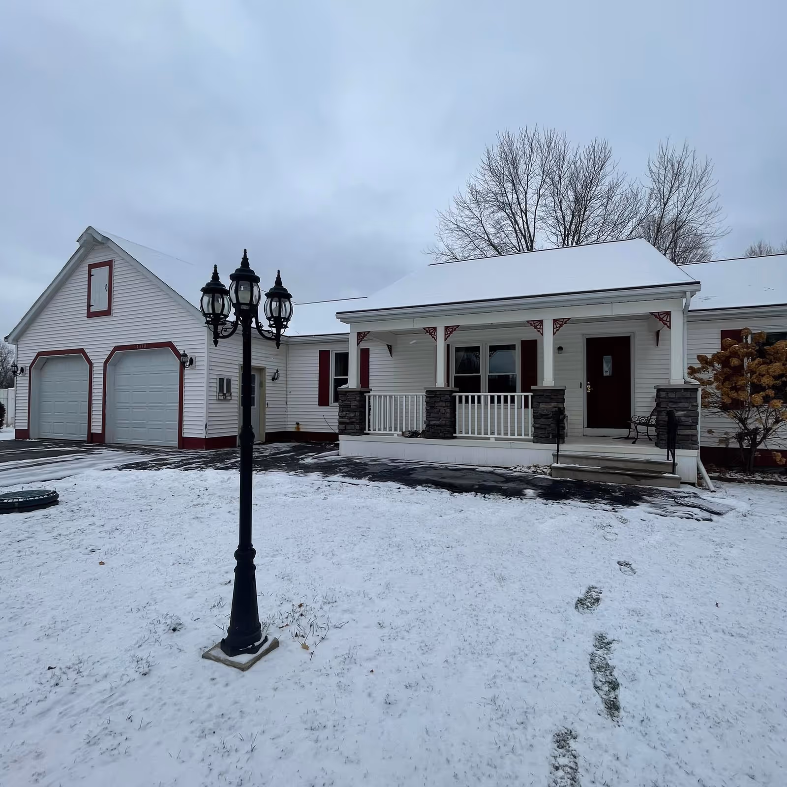 Snow-covered front yard and driveway leading to a white single-story house with a covered porch, attached two-car garage, and a black lamppost.