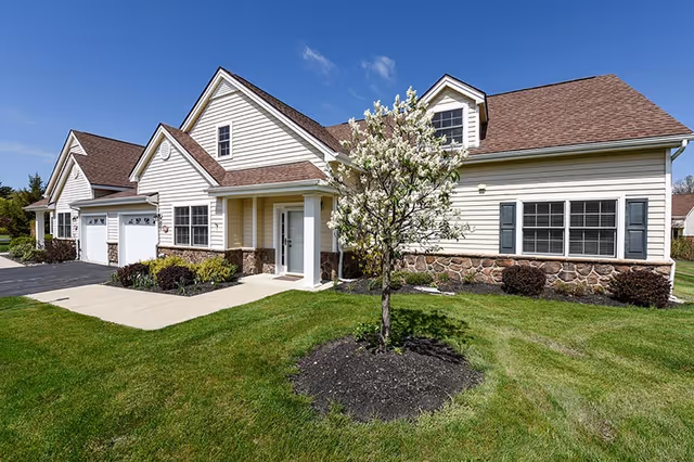 Exterior view of a single-story residential building with beige siding, stone accents, and a brown shingled roof. There is a small tree with white blossoms in the front yard, green grass, and a concrete walkway leading to the front door. The sky is clear and blue.