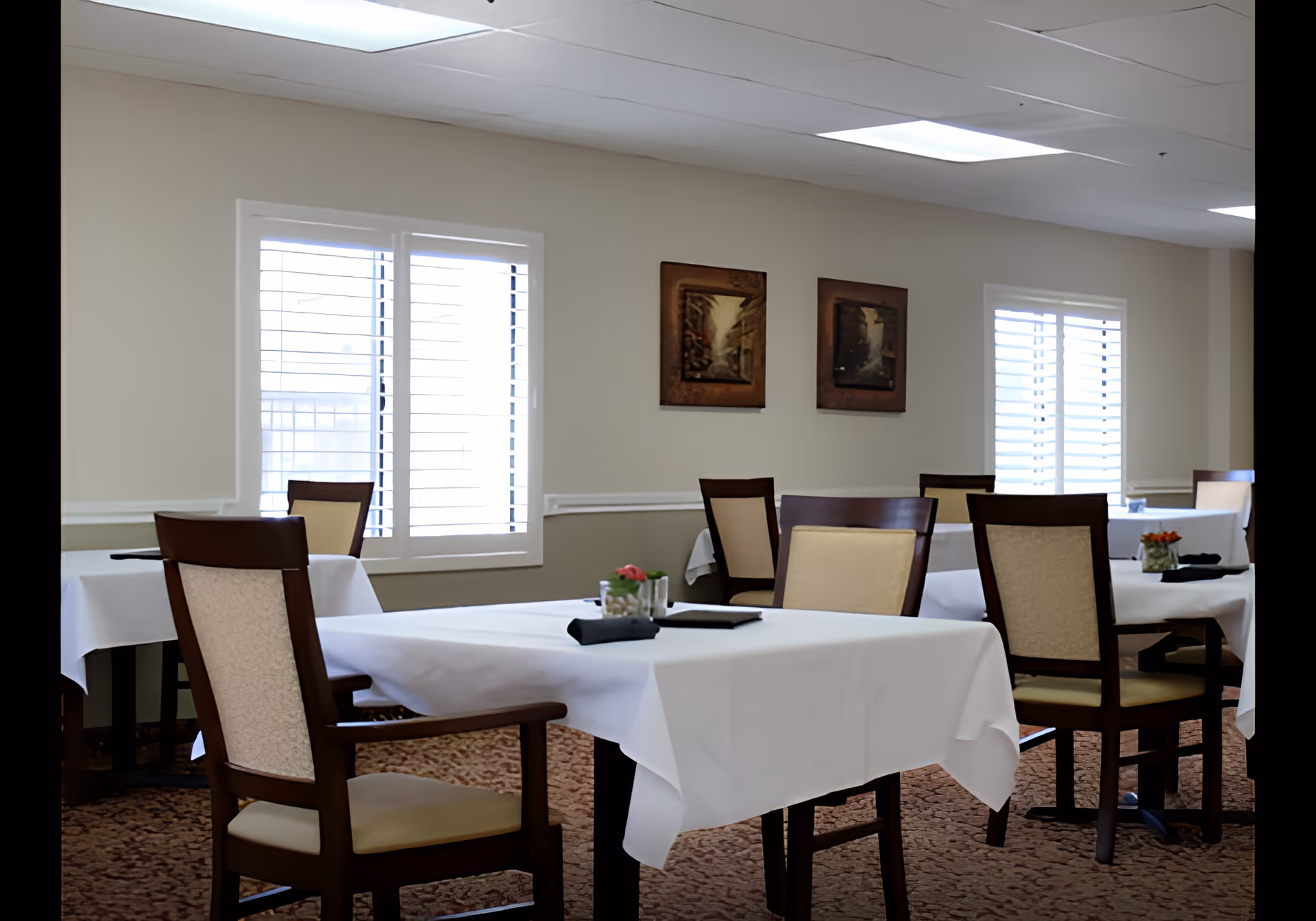 A dining room with several tables covered in white tablecloths, each surrounded by wooden chairs with beige cushions. The room has beige walls with two framed pictures hanging and windows with white shutters allowing natural light to enter. The floor is carpeted with a patterned design.