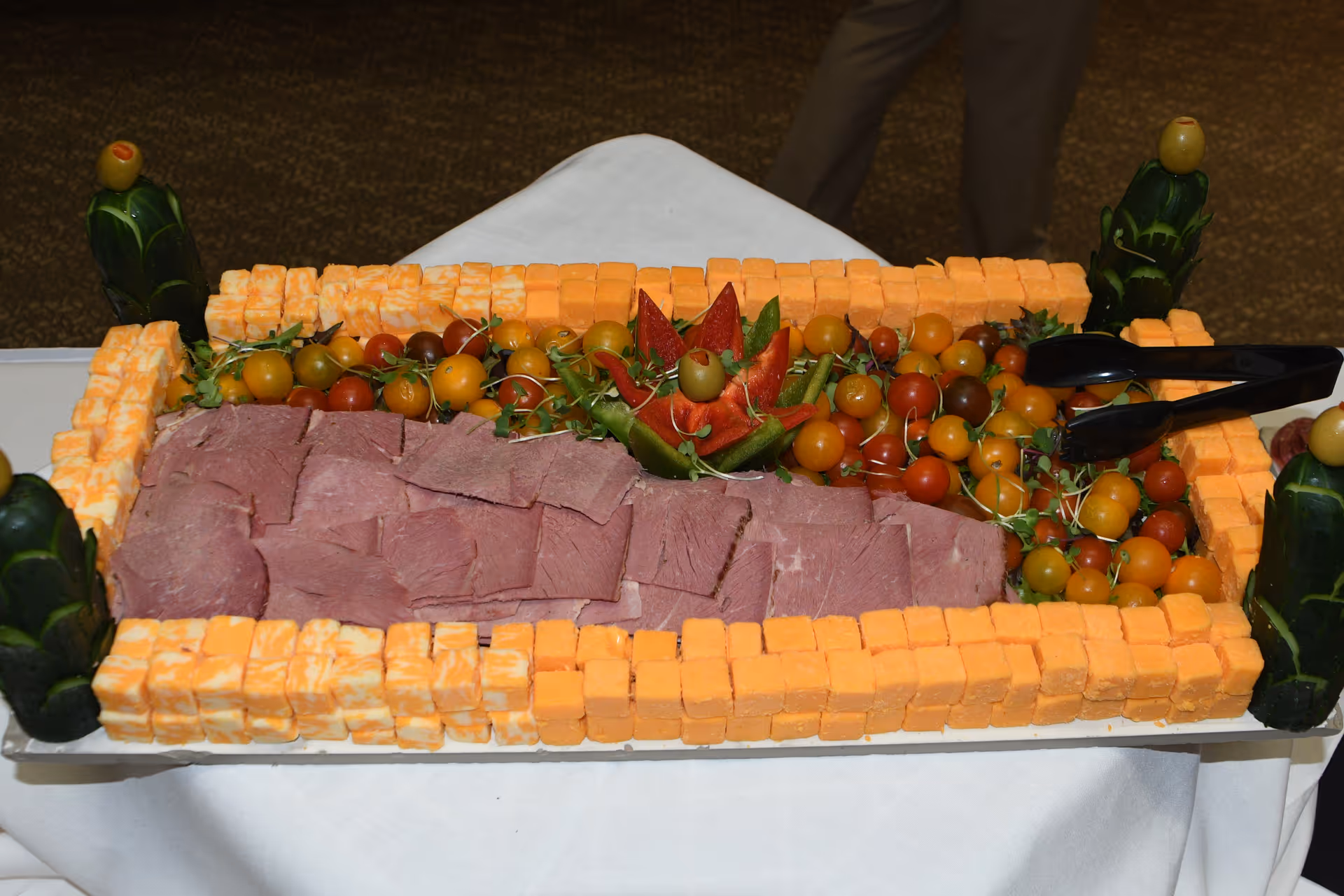 A rectangular platter of sliced roast beef surrounded by cubes of cheddar and marbled cheese, cherry tomatoes and decorative carved vegetables on a white tablecloth.