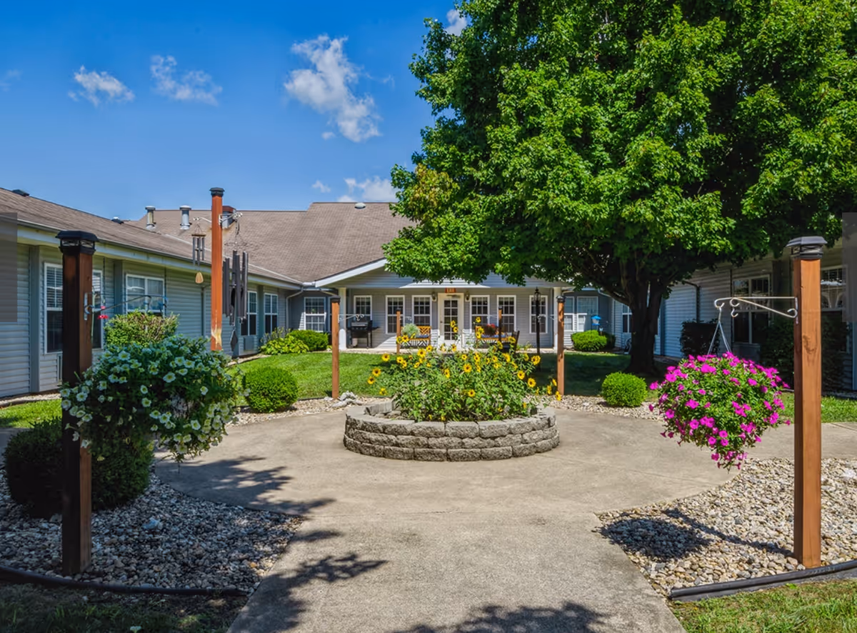 Outdoor courtyard area at Cedar Creek of Logansport featuring a circular flower bed with sunflowers in the center, surrounded by a concrete walkway. There are hanging flower baskets on wooden posts, green bushes, and a large leafy tree providing shade. The courtyard is enclosed by single-story building wings with multiple windows and a clear blue sky above.