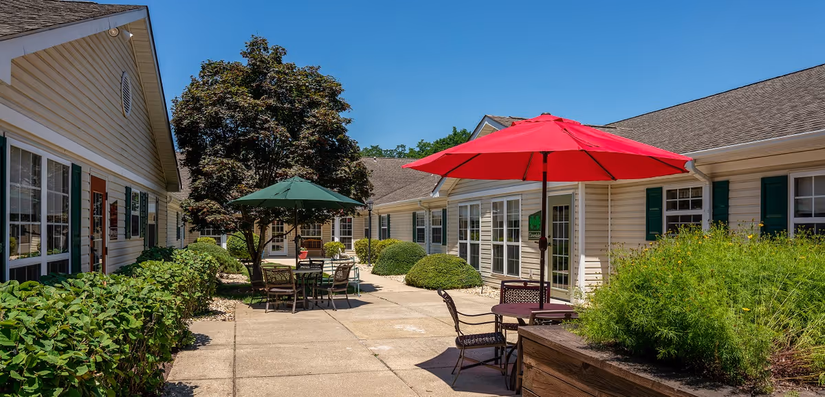 Sunny courtyard with patio tables, red and green umbrellas, and landscaping between single-story facility buildings.