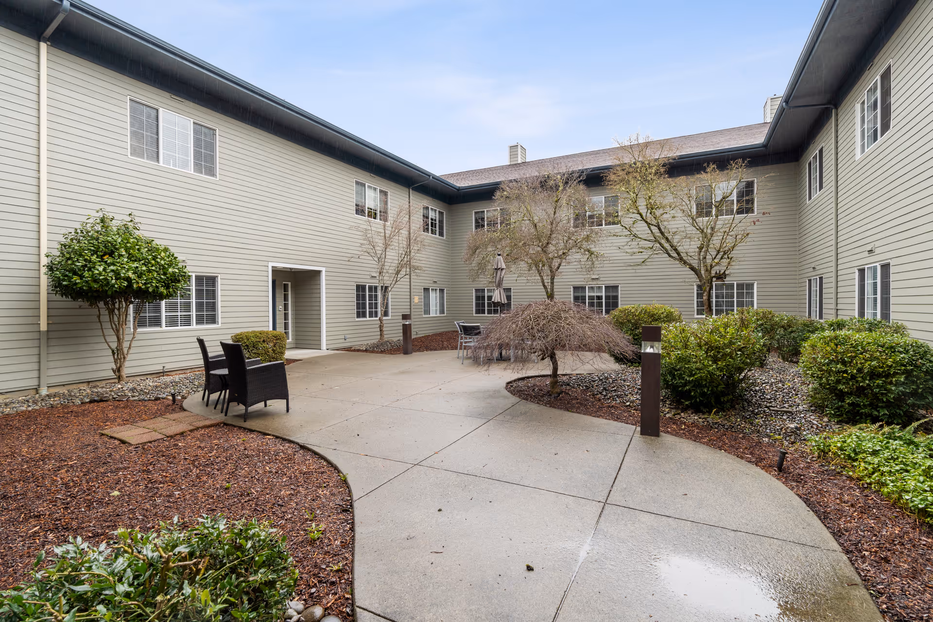 Outdoor courtyard of a two-story assisted living building with a paved walkway, patio seating, trees, and landscaping.