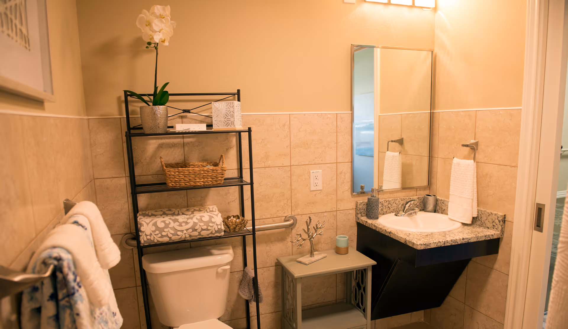 Bathroom with a toilet, metal shelving unit, wall-mounted sink and mirror, and decorative towels and accessories.