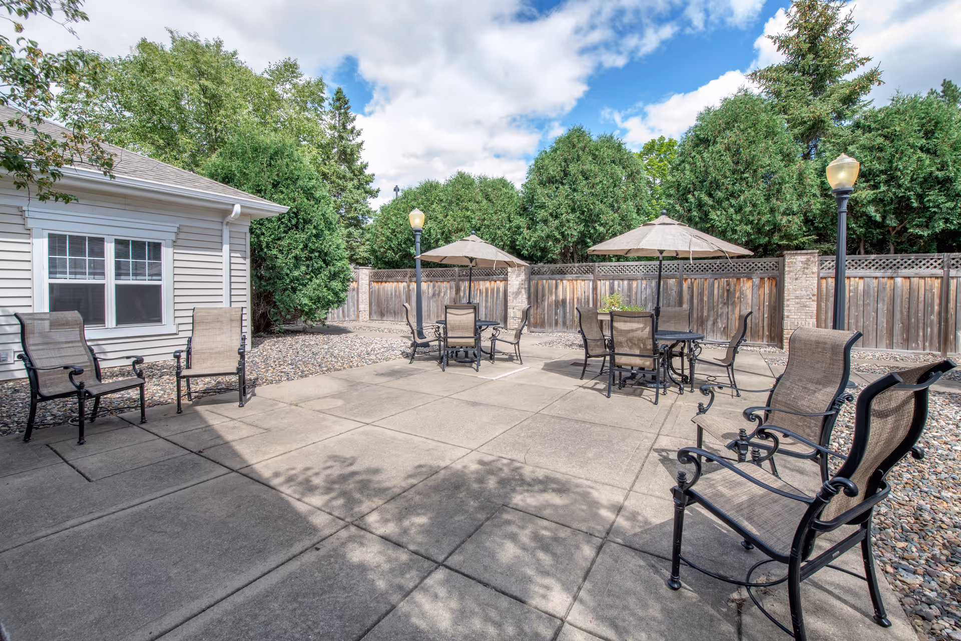 Outdoor patio area with several metal chairs and tables with umbrellas, surrounded by a wooden fence and tall green trees under a partly cloudy sky.