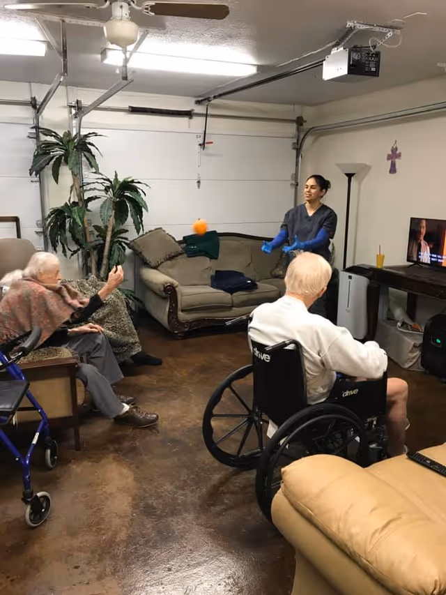 A group of elderly people and a caregiver are in a living room setting. Two elderly individuals are seated, one in a wheelchair and another on a chair with a walker nearby. The caregiver is standing and appears to be engaging with them by tossing an orange ball. The room has a couch, a TV on a stand, a floor lamp, and some indoor plants. The floor is a polished brown surface and the walls are white with a garage door visible in the background.