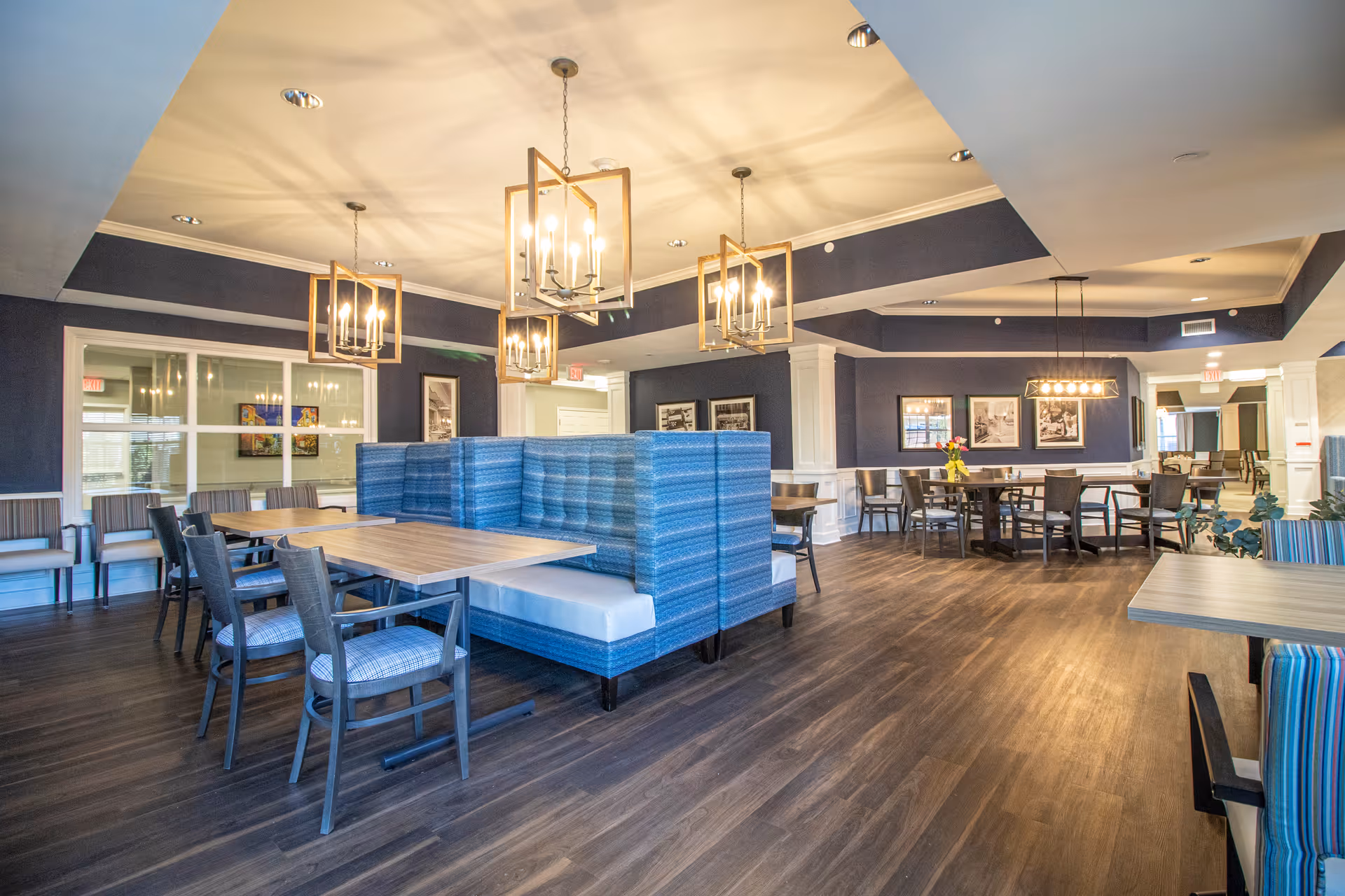 A spacious dining room with wooden floors, multiple tables and chairs, and blue upholstered booth seating. The walls are painted dark blue with white trim, and there are several modern chandeliers hanging from the ceiling. Framed black and white photographs decorate the walls.