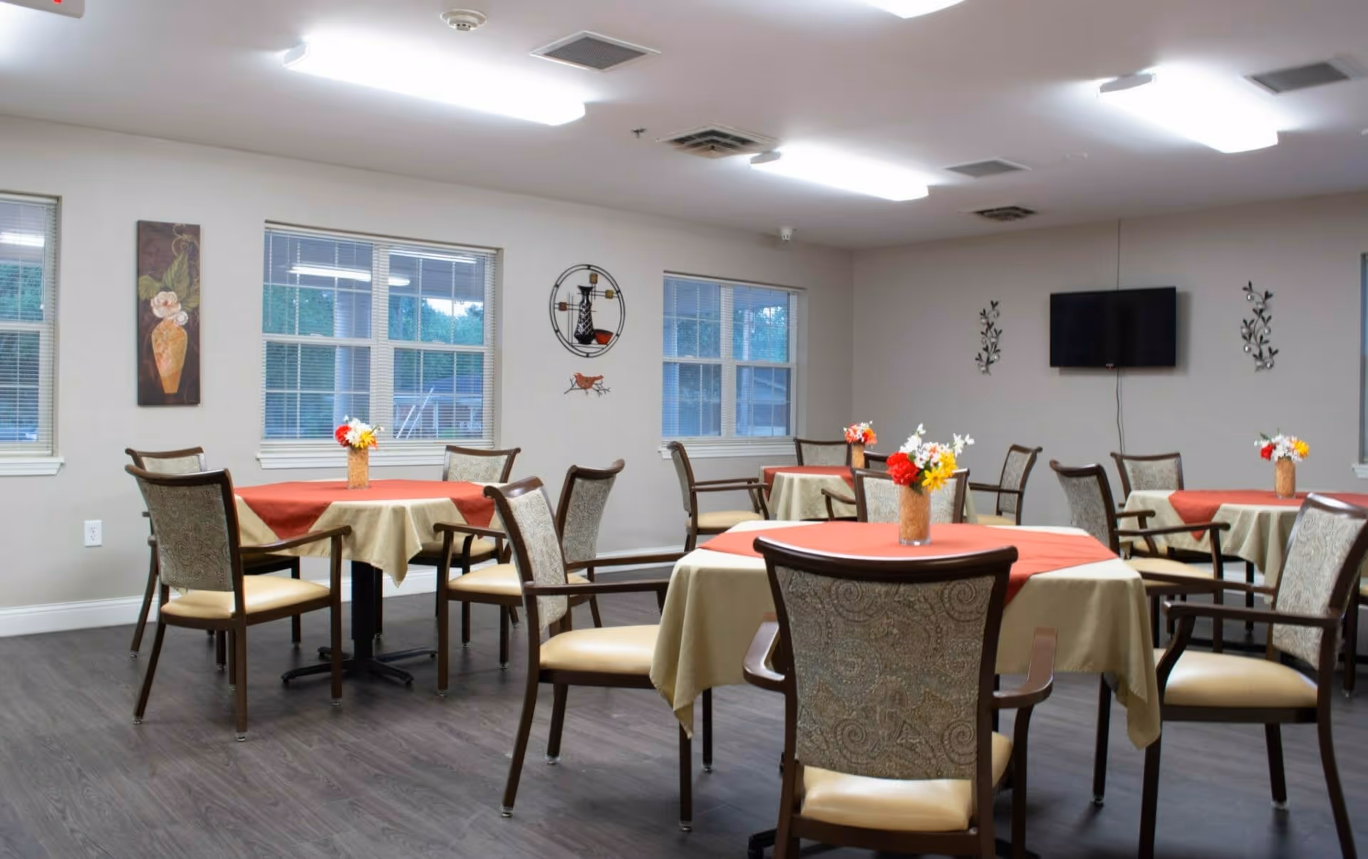 A dining room with several round tables covered with beige and red tablecloths, each table decorated with a small vase of colorful flowers. The room has multiple windows with blinds, wall decorations including a painting and metal art, a mounted flat-screen TV, and overhead fluorescent lighting. The floor is dark wood.