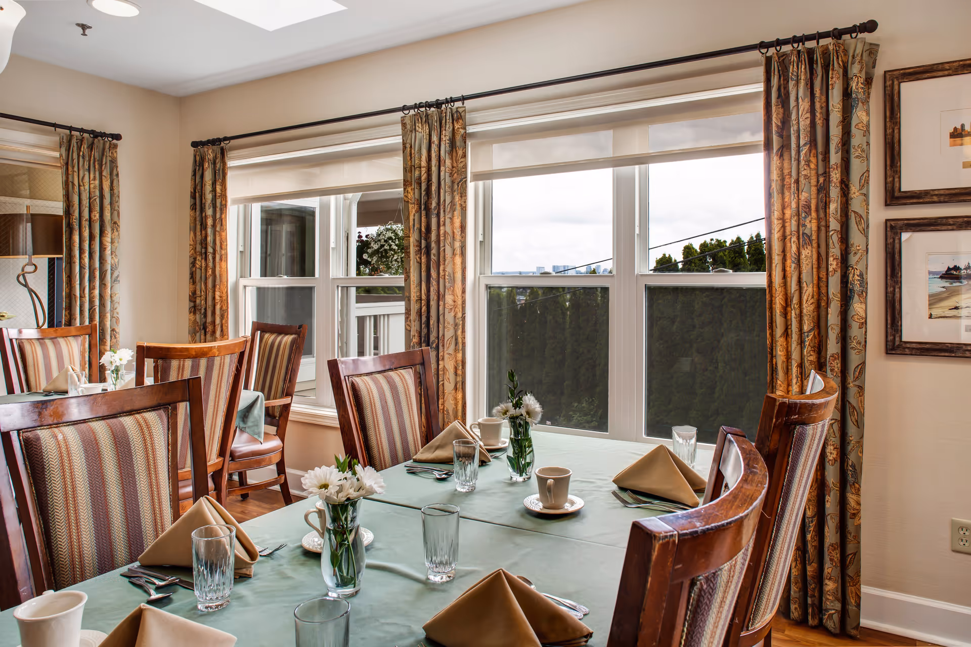 A dining room with a table set for a meal, featuring green tablecloth, beige folded napkins, clear drinking glasses, white coffee cups on saucers, and small vases with white flowers. The room has large windows with floral curtains, wooden chairs with striped upholstery, and framed artwork on the wall.