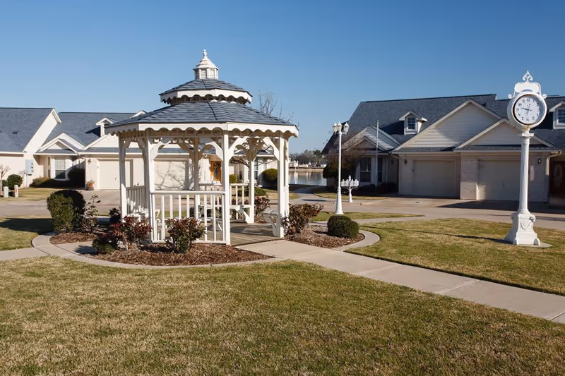 A white gazebo with a shingled roof stands in the center of a well-maintained grassy courtyard with paved walkways. Surrounding the gazebo are small bushes and landscaped mulch beds. In the background, there are single-story residential buildings with garages and a decorative white clock on a post. The sky is clear and blue.