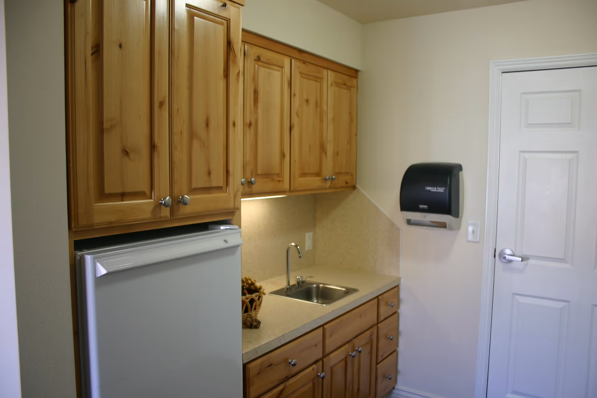 Small kitchenette area with wooden cabinets, a countertop with a small sink, a white mini refrigerator, and a black paper towel dispenser mounted on the wall next to a white door.