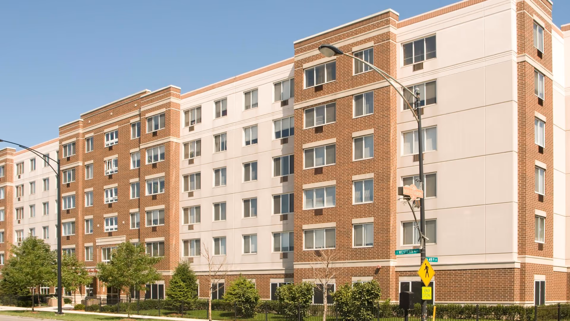 Exterior view of a multi-story senior living facility building with a combination of brick and light-colored paneling. The building has many windows and is surrounded by small trees and shrubs along the sidewalk. A street sign and pedestrian crossing sign are visible in front of the building under a clear blue sky.