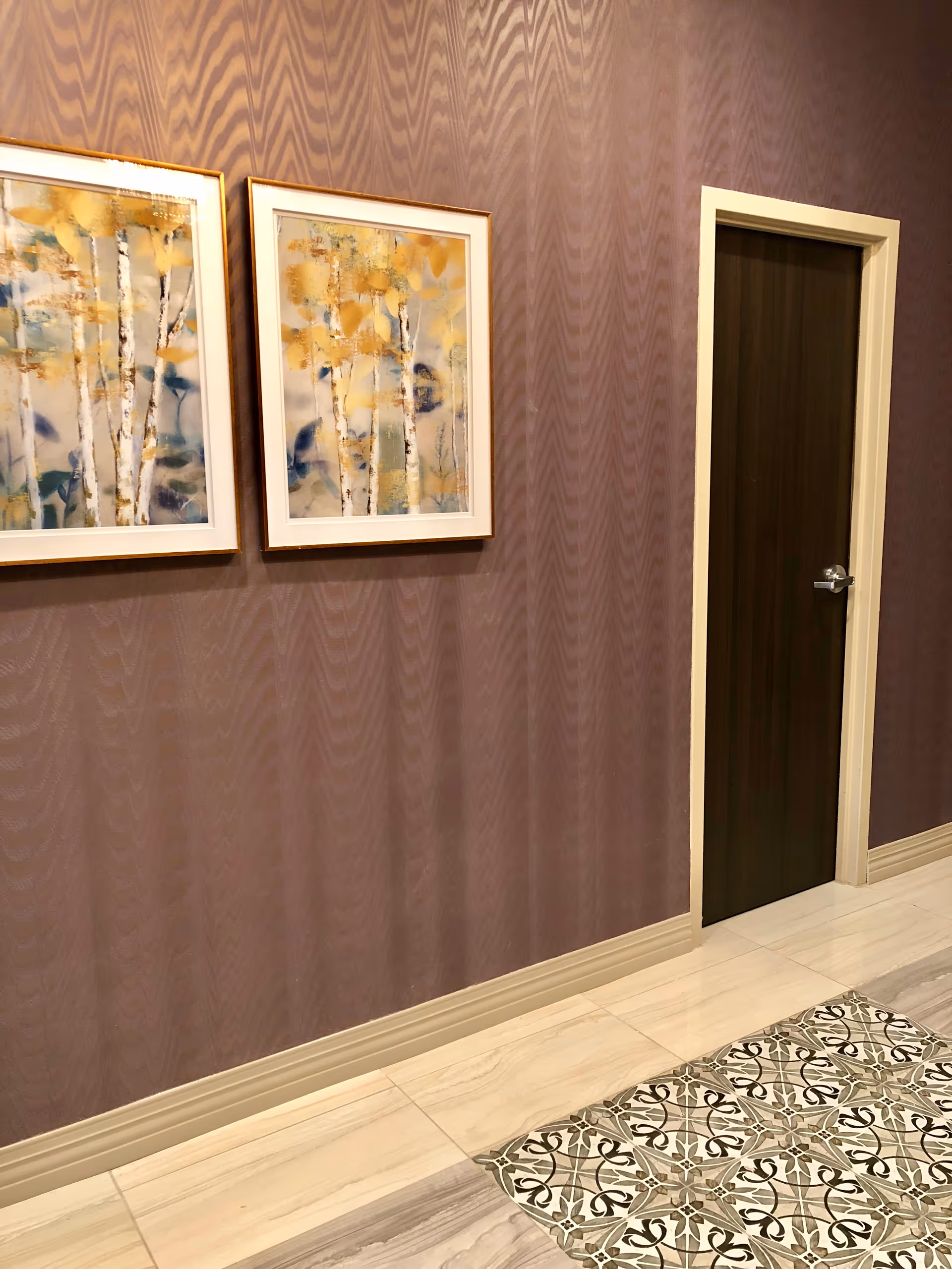 Interior hallway with purple textured wallpaper, two framed abstract paintings featuring birch trees with yellow and blue accents, a dark wooden door with a silver handle, and a floor with beige tiles and a decorative patterned tile section.