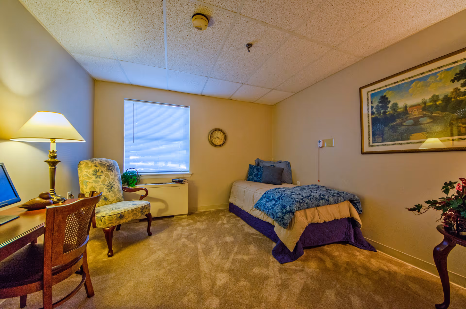 A cozy bedroom in a senior living facility featuring a single bed with blue and beige bedding, a floral upholstered armchair, a wooden desk with a chair, a table lamp, a window with blinds, a wall clock, a framed landscape painting, and a small table with a plant.