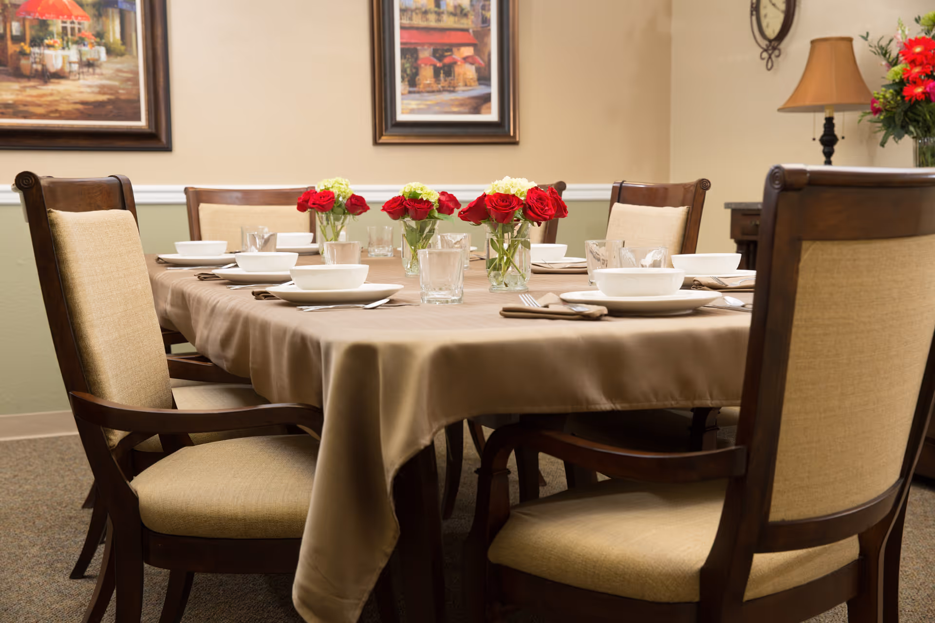 A dining room table set for a meal with beige tablecloth, white bowls and plates, glasses, and silverware. There are three small vases with red and white flowers as centerpieces. The room has beige walls with two framed paintings, a clock, a lamp, and a flower arrangement on a side table.