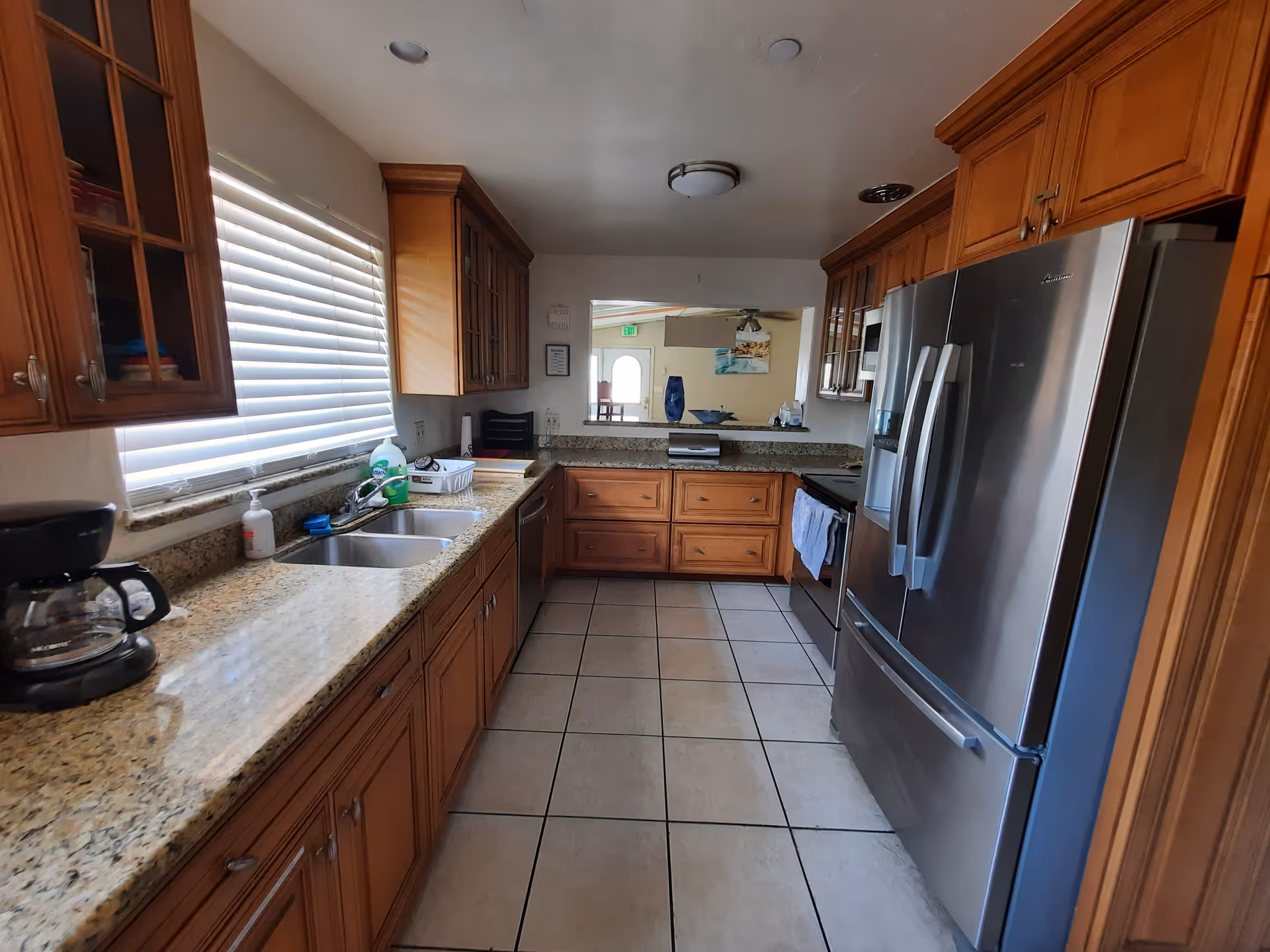 A kitchen with wooden cabinets, granite countertops, a double sink, a coffee maker, a stainless steel refrigerator, and a stove. There is a window with blinds on the left side and a pass-through opening to another room at the far end.
