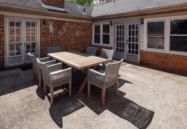 Outdoor patio area with a wooden table and six wicker chairs arranged around it, surrounded by brick walls with multiple windows and doors leading inside the building.