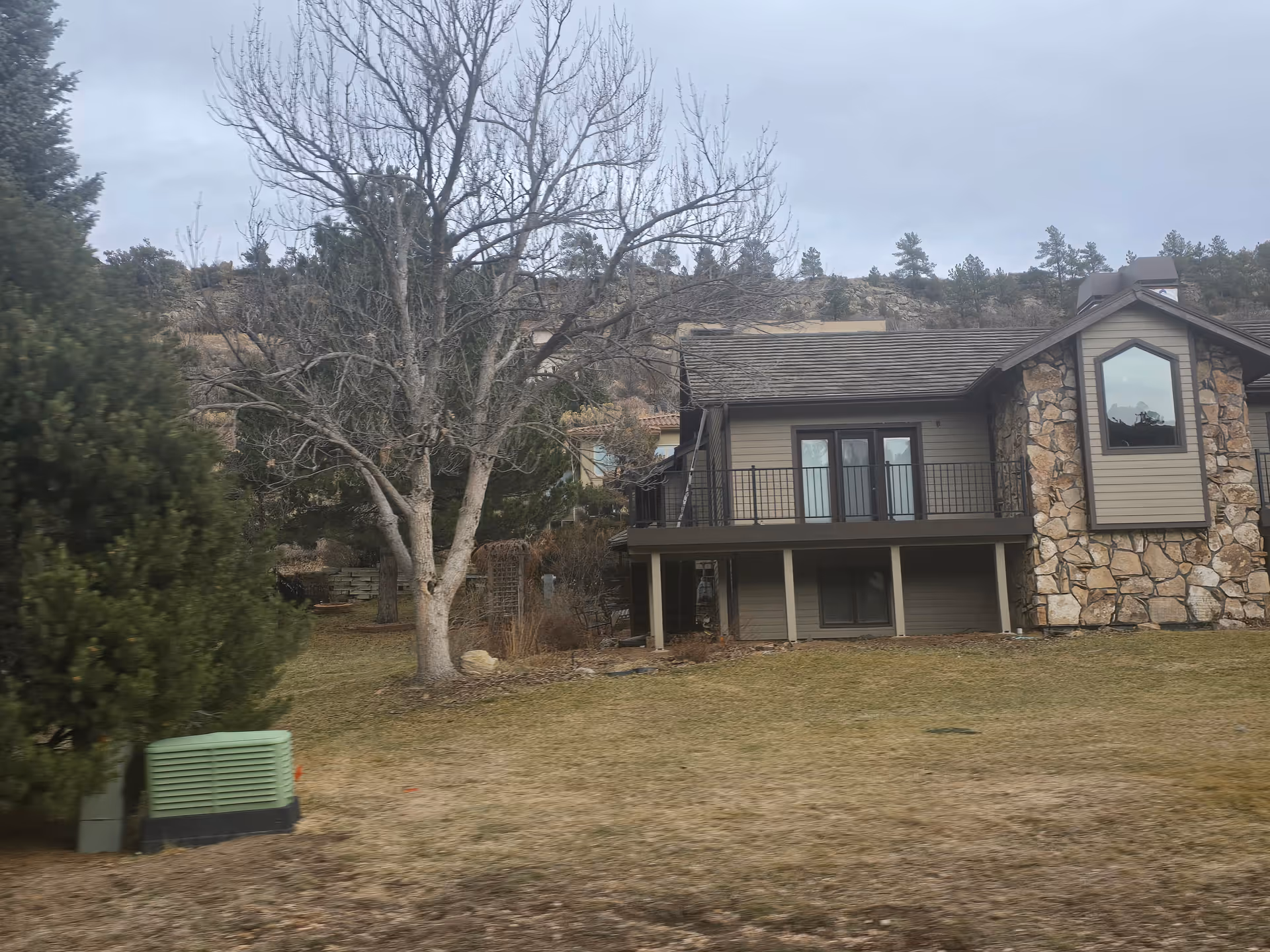 Exterior view of a house with stone and wood siding, a balcony with black railing, and a large window. The yard has a mix of grass and bare patches, with several trees including a leafless tree in the foreground. Hills and trees are visible in the background under a cloudy sky.