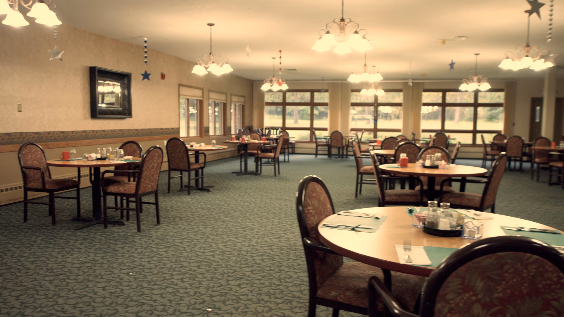Spacious dining room with round tables and chairs set for meals and large windows along the back wall.