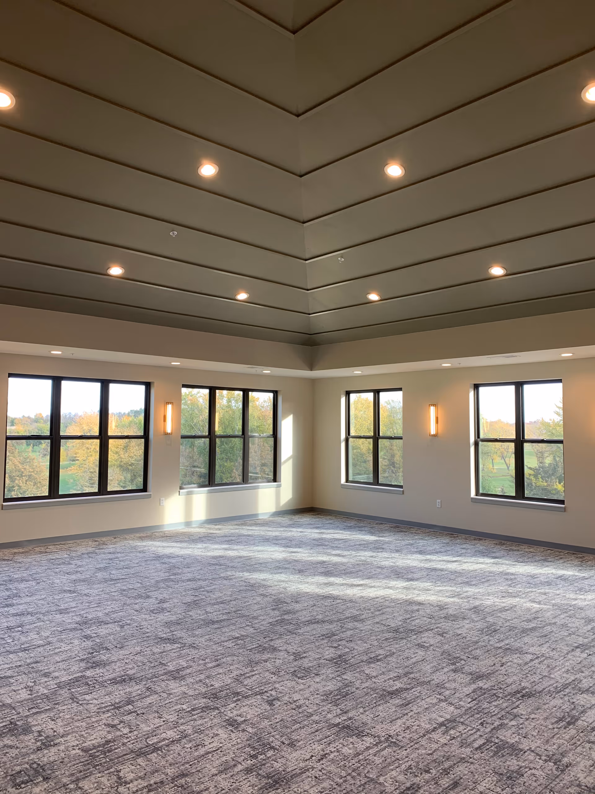 A spacious, empty room with large windows on two walls allowing natural light to fill the space. The ceiling is high with recessed lighting and a layered design. The floor is carpeted with a textured pattern, and the walls are painted in a neutral color with modern wall sconces.