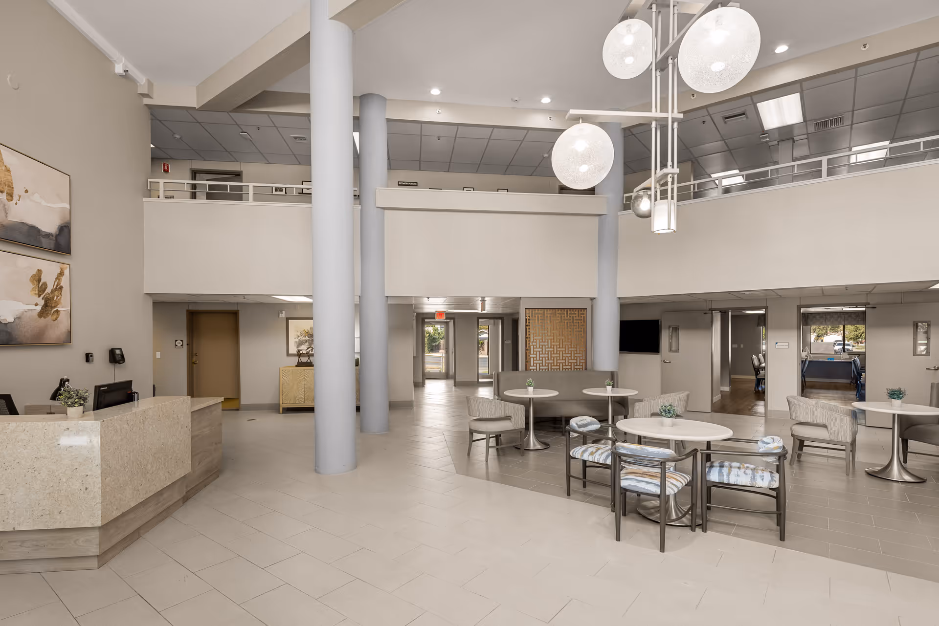 Spacious and modern senior living community lobby with high ceilings, large round pendant lights, a reception desk on the left, and several seating areas with tables and chairs. The area features neutral tones and decorative wall art, with a second-floor balcony overlooking the lobby.