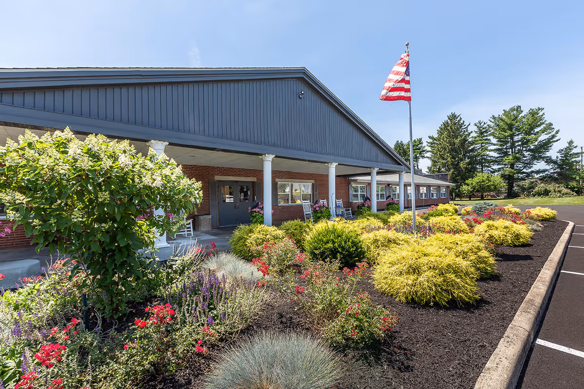 Exterior view of Continuing Healthcare of Milan building with a covered entrance, an American flag on a flagpole, and a landscaped garden with various shrubs and flowers in front. The sky is clear and blue.