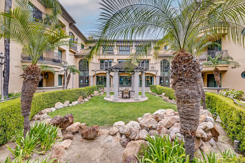 Outdoor courtyard area at Meridian at Chino featuring a circular gazebo with columns and seating in the center, surrounded by green grass, palm trees, trimmed hedges, rocks, and a multi-story building with balconies in the background.