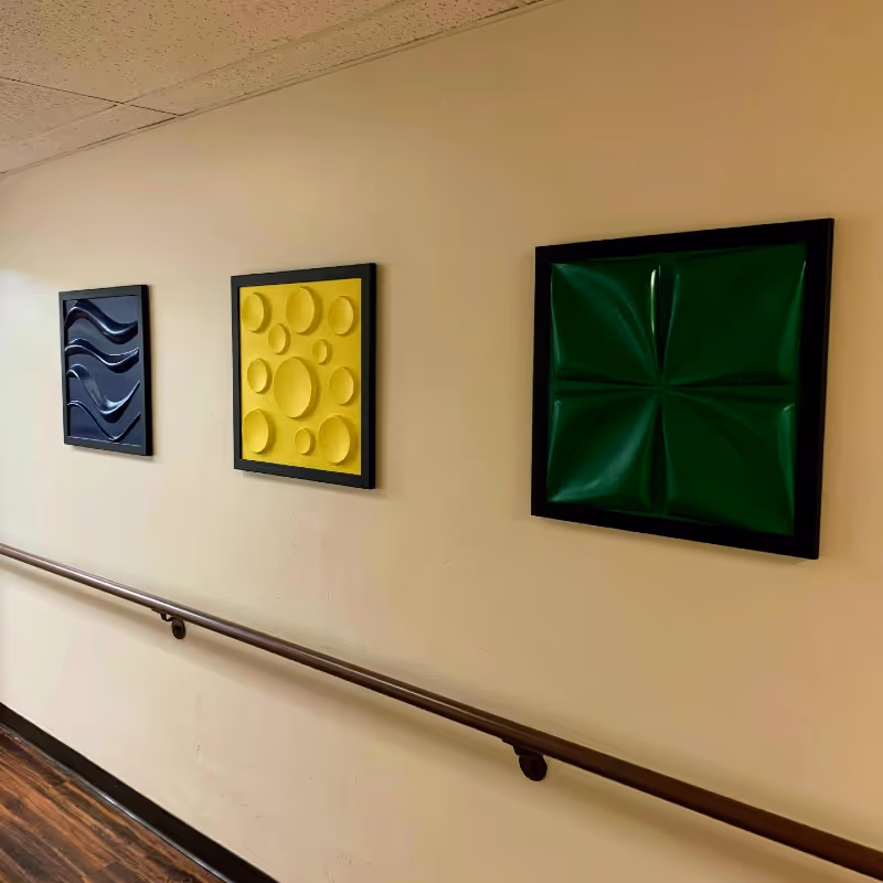 A beige hallway wall with three square framed abstract art pieces in black frames. The art pieces feature textured designs in black, yellow, and green. A wooden handrail runs along the wall below the artwork.