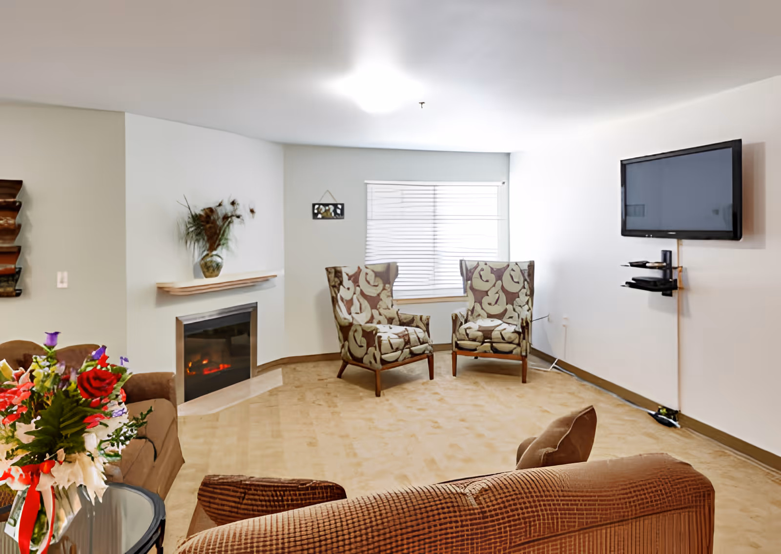 Bright living room with two patterned armchairs by a window, wall-mounted TV, fireplace, sofas, and a floral arrangement on a side table.