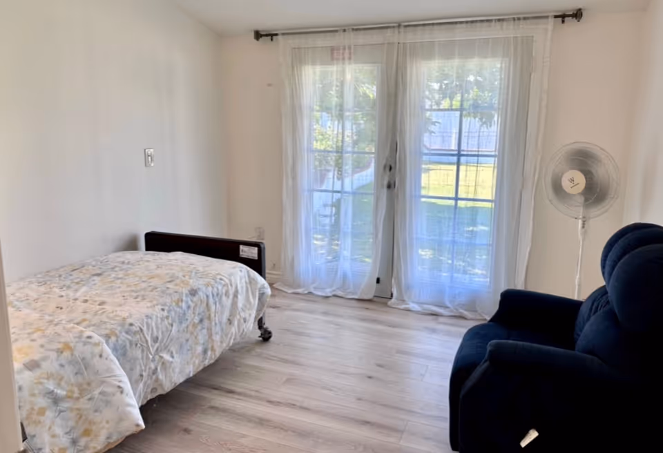 A small bedroom with a single bed, sheer curtains, a standing fan, and a blue recliner