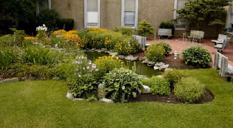 A landscaped garden area with green grass, various flowering plants, and bushes surrounding a small pond. There are stone borders around the pond and several benches and chairs on a brick patio area near the building in the background.