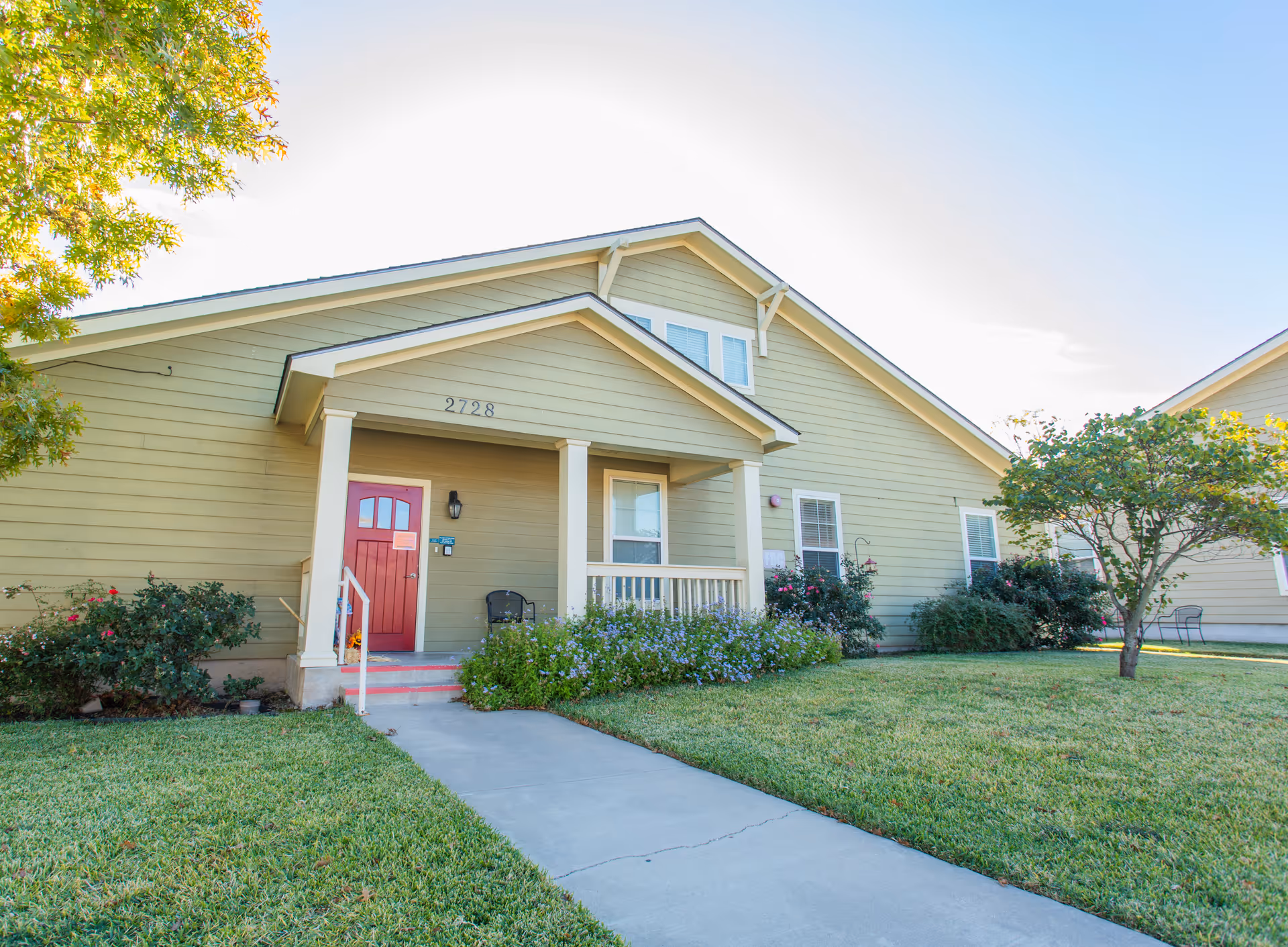 Exterior view of a single-story building with beige siding and a red front door numbered 2728. There is a small porch with white columns, a chair, and a well-maintained lawn with bushes and a tree. The sky is clear and blue.