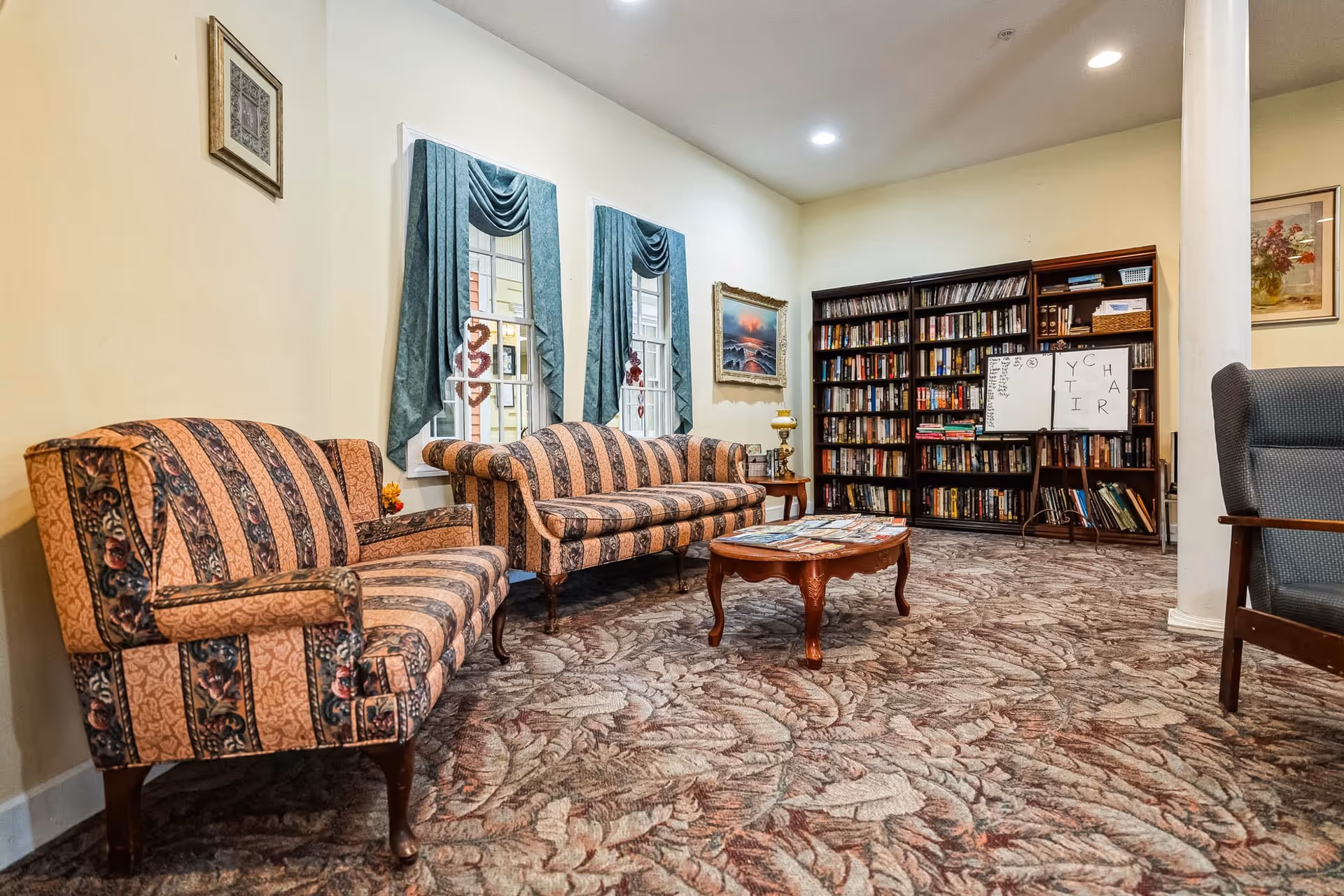 A cozy living room area with two vintage-style striped sofas, a wooden coffee table with magazines, a large bookshelf filled with books, and a whiteboard with letters and words. The room has patterned carpet, cream-colored walls, teal curtains on two windows, and framed artwork on the walls.