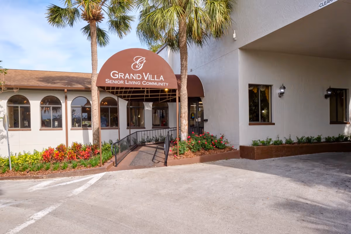 Exterior entrance of Grand Villa Senior Living Community with a brown awning displaying the facility name. The entrance is flanked by palm trees and landscaped flower beds with colorful plants. The building has white walls and several windows, and there is a paved driveway in front.