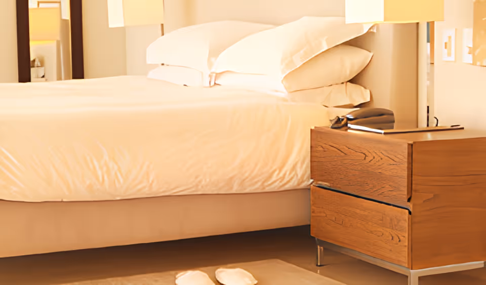 A neatly made bed with white linens and multiple pillows in a softly lit bedroom. Next to the bed is a wooden nightstand with two drawers, holding a telephone and a closed book or folder. A pair of white slippers is placed on the floor beside the bed.