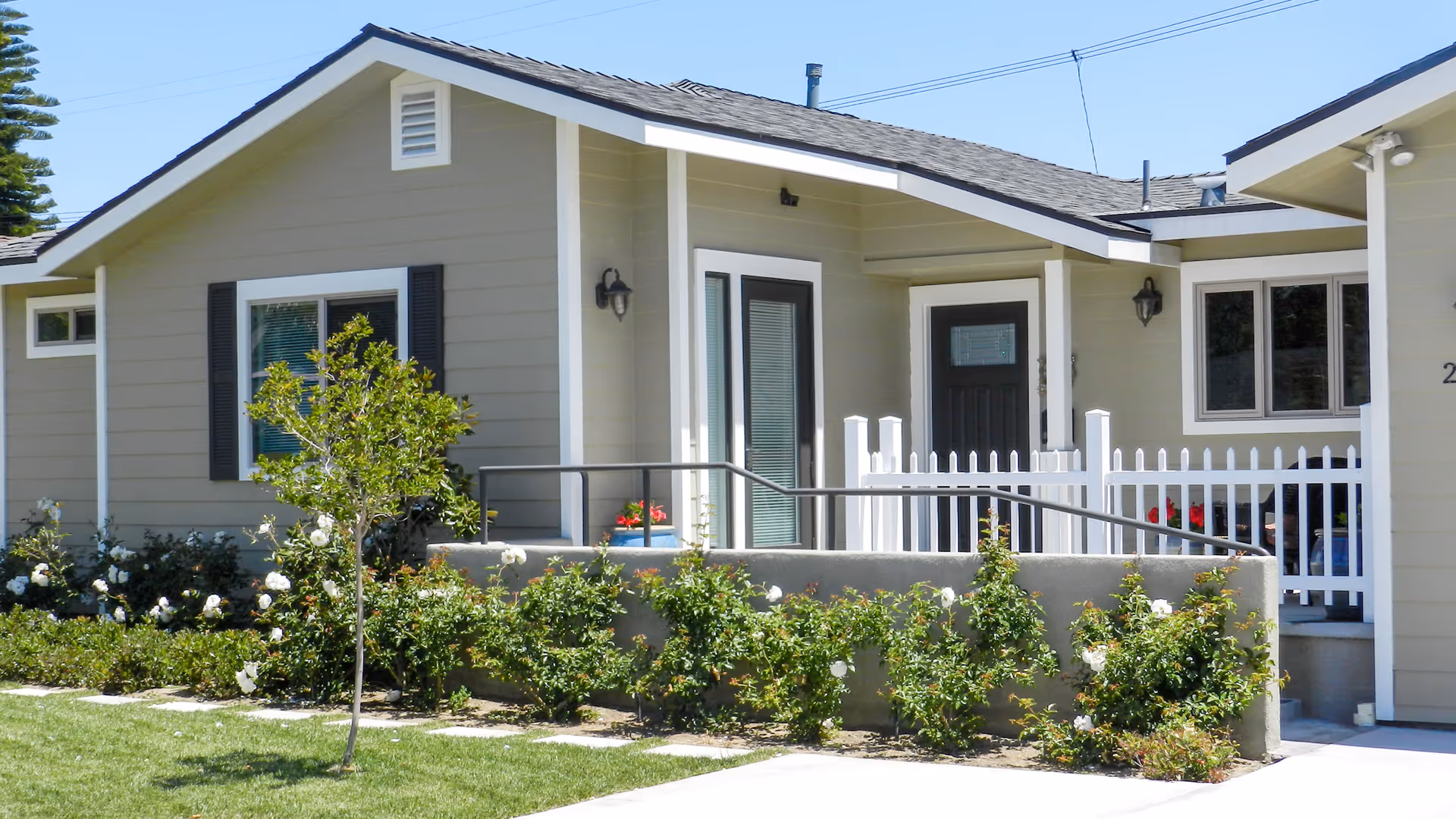 Exterior view of a single-story residential building with beige siding, white trim, and a dark roof. The entrance features a black door and a small porch area with a white picket fence and a ramp with handrails. There are bushes with white flowers and a small tree in front of the building, along with a well-maintained lawn.