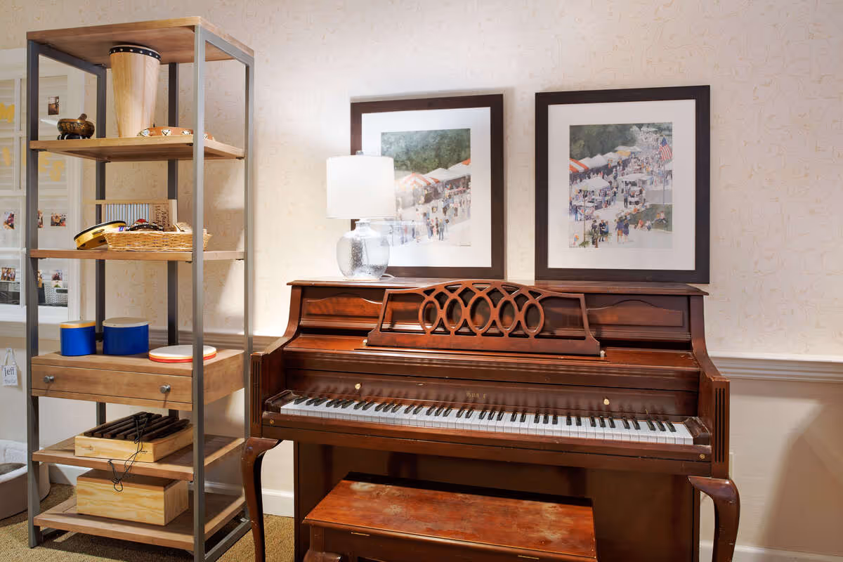 A wooden upright piano with a matching bench in front of a beige patterned wall. On top of the piano is a glass lamp with a white shade. Above the piano are two framed pictures depicting outdoor scenes with people and tents. To the left of the piano is a tall shelving unit holding various musical instruments and items, including drums, tambourines, and a wooden box.
