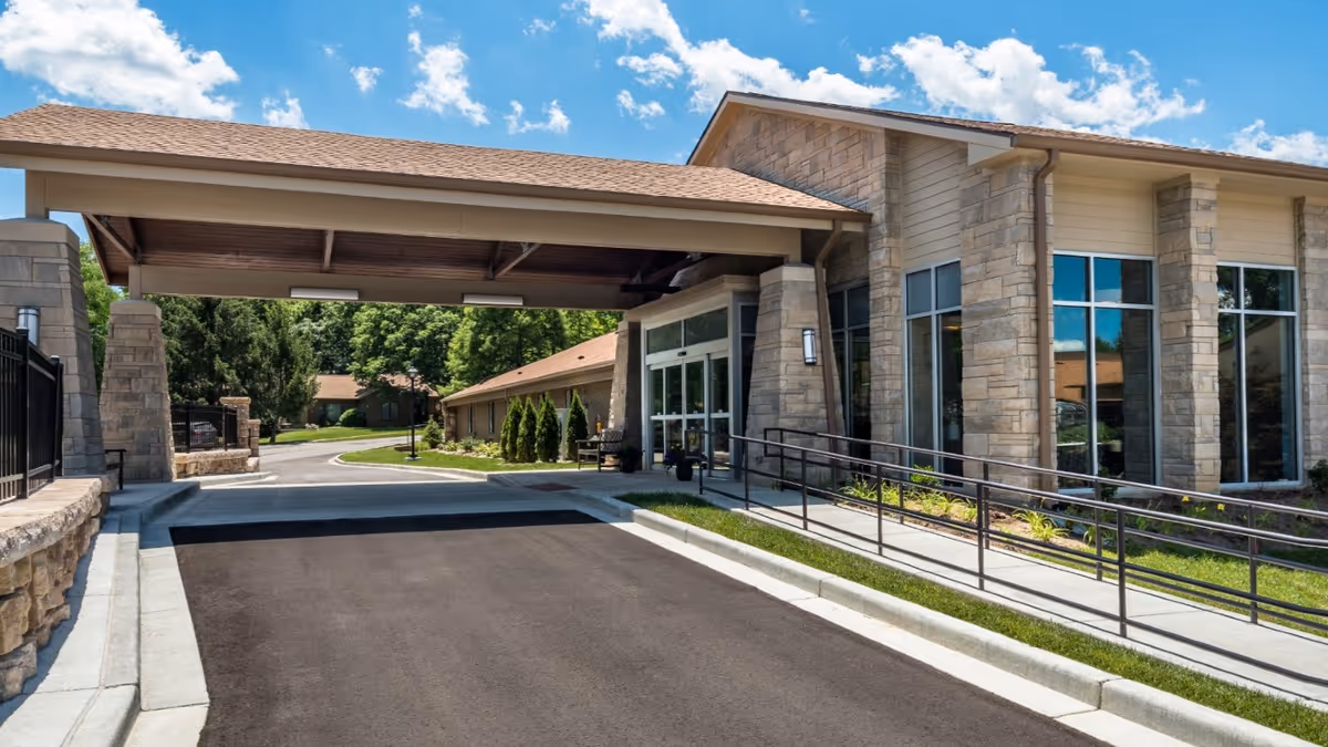 Entrance of a senior living facility named Meadowood with a covered drop-off area, stone pillars, large windows, a wheelchair accessible ramp, and a driveway leading into a green landscaped area with trees under a blue sky with clouds.