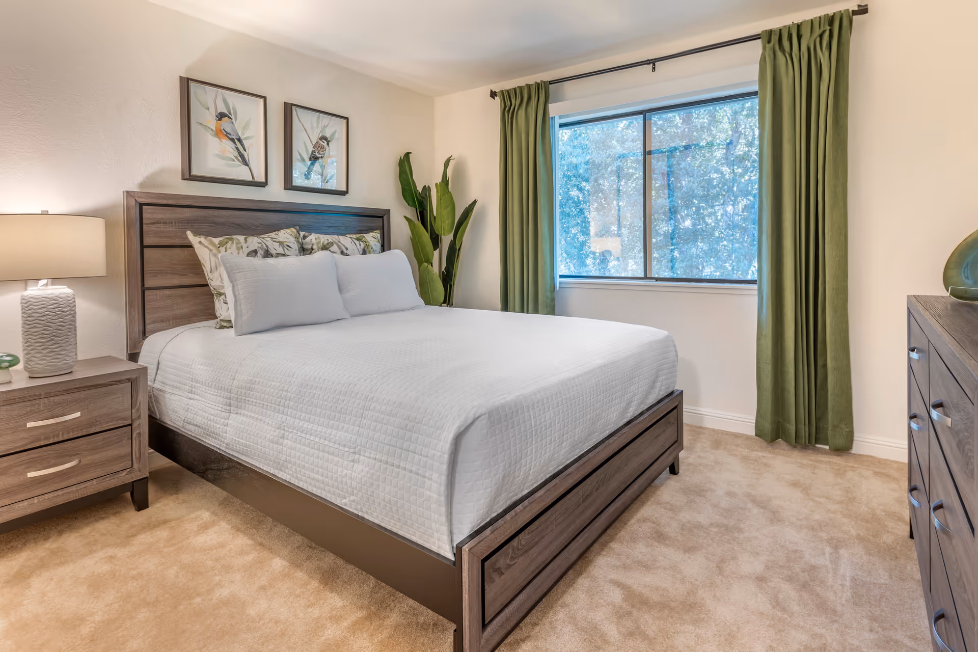 A bedroom with a large bed featuring white bedding and floral pillows, a wooden headboard, a nightstand with a lamp, a dresser, green curtains on a window showing trees outside, and two framed bird pictures on the wall.