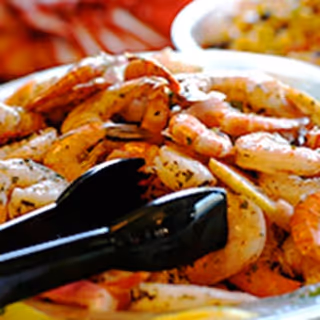 Close-up of a plate filled with cooked shrimp seasoned with herbs, with black tongs resting on top and a blurred background showing another dish.