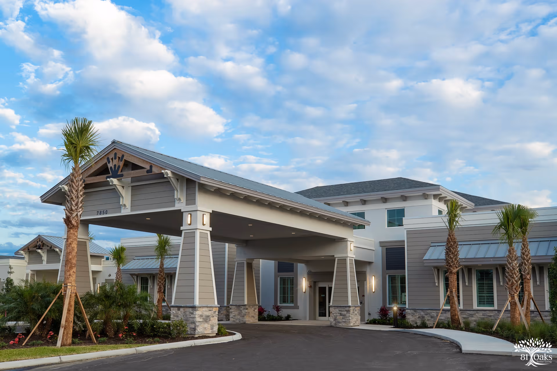 Exterior view of 81 Oaks Senior Living facility showing a covered entrance with stone pillars, palm trees, and a clear sky with scattered clouds.
