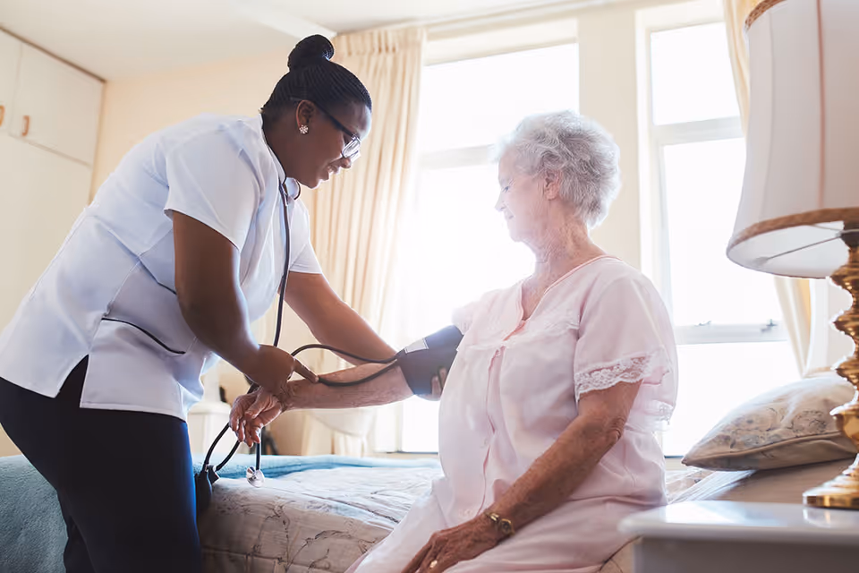 A healthcare worker measures an elderly woman's blood pressure while she sits on a bed in a bright bedroom.