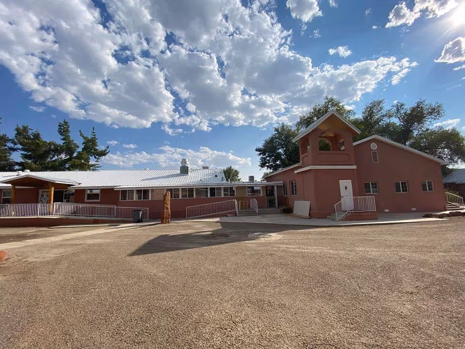 Exterior view of Casa de Rosa Assisted Living facility showing a single-story building with a light-colored roof and a small tower-like structure. The building is surrounded by trees under a partly cloudy blue sky. There is a paved area in front of the building with ramps and stairs leading to the entrances.