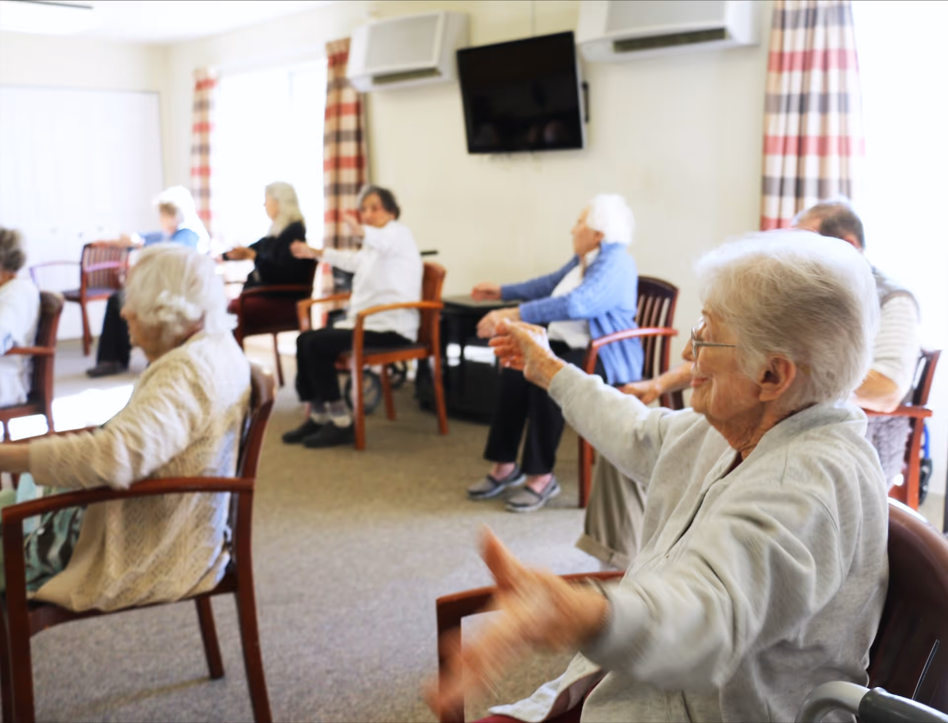 Several elderly residents seated in a communal activity room doing seated arm exercises.