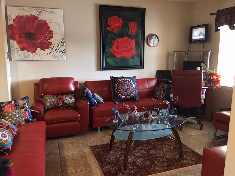 Cozy living room with red leather sofas and armchairs, a glass coffee table displaying decorative glass sculptures, colorful throw pillows, and floral wall art.