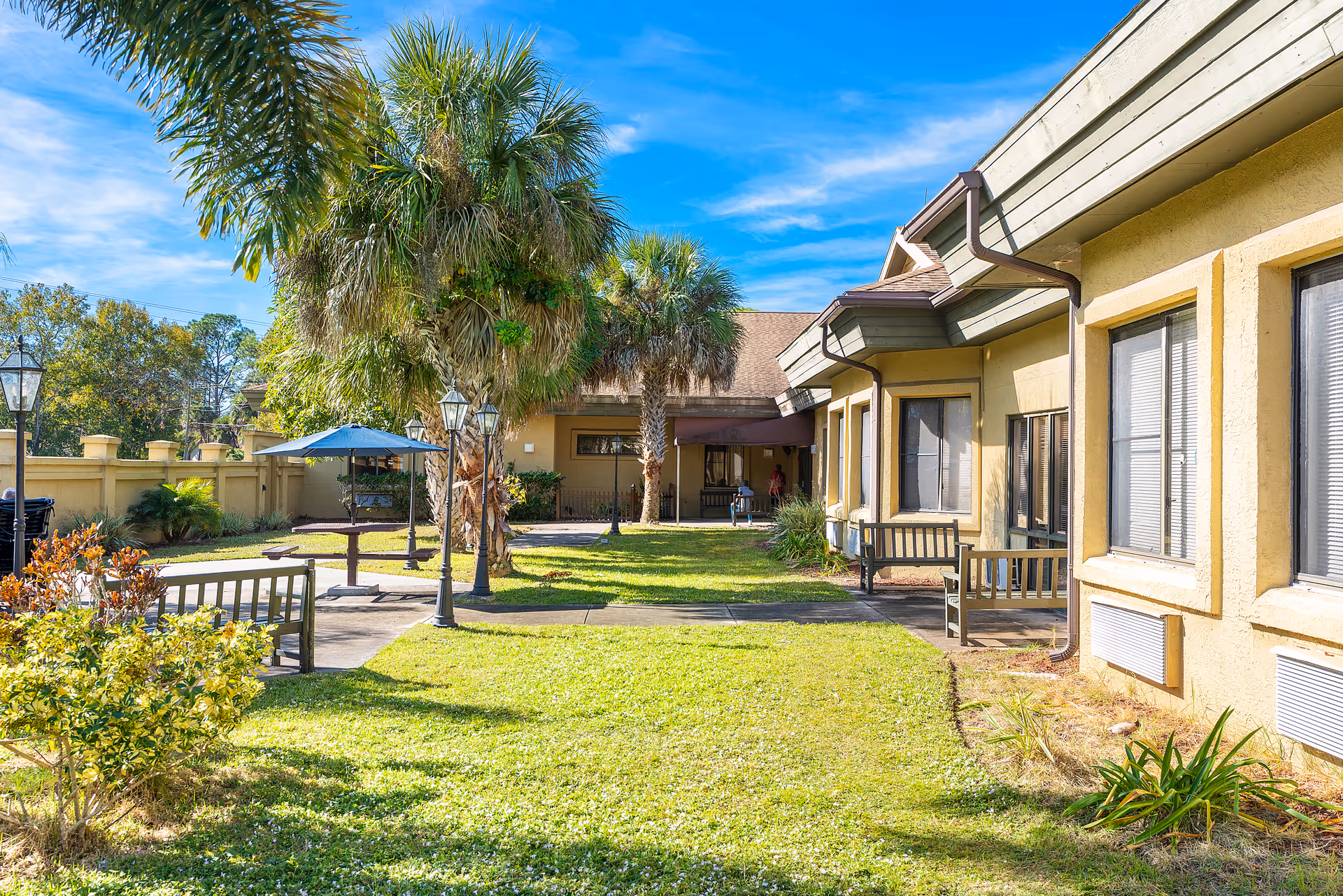 Outdoor courtyard area with green grass, palm trees, benches, and a table with an umbrella. The courtyard is surrounded by a beige building with windows and a fence, under a bright blue sky with some clouds.