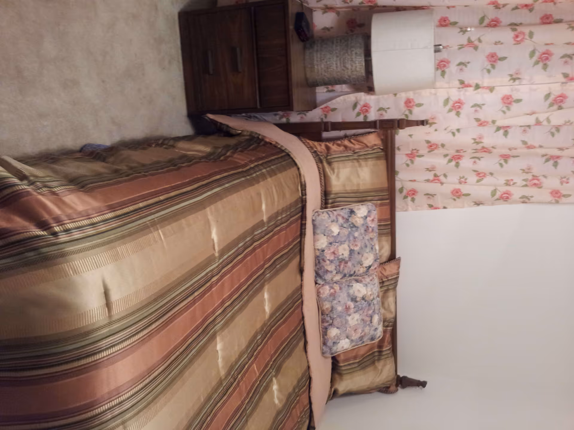 A bedroom with a bed covered in a striped brown, beige, and green bedspread. Two floral pillows are placed on the bed. Next to the bed is a wooden nightstand with a white lamp and a decorative basket. The wall behind the bed has floral curtains with a pink rose pattern.