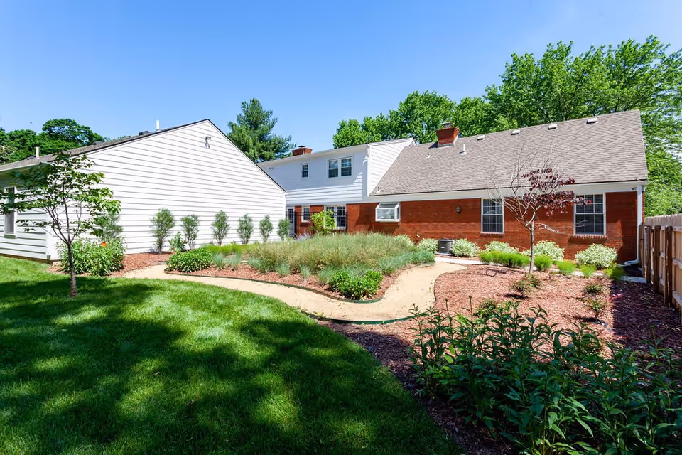 A sunny outdoor garden area with a curved gravel pathway surrounded by green grass, shrubs, and small trees. The garden is bordered by a white siding building on the left and a red brick building with a gray roof on the right. The sky is clear and blue.