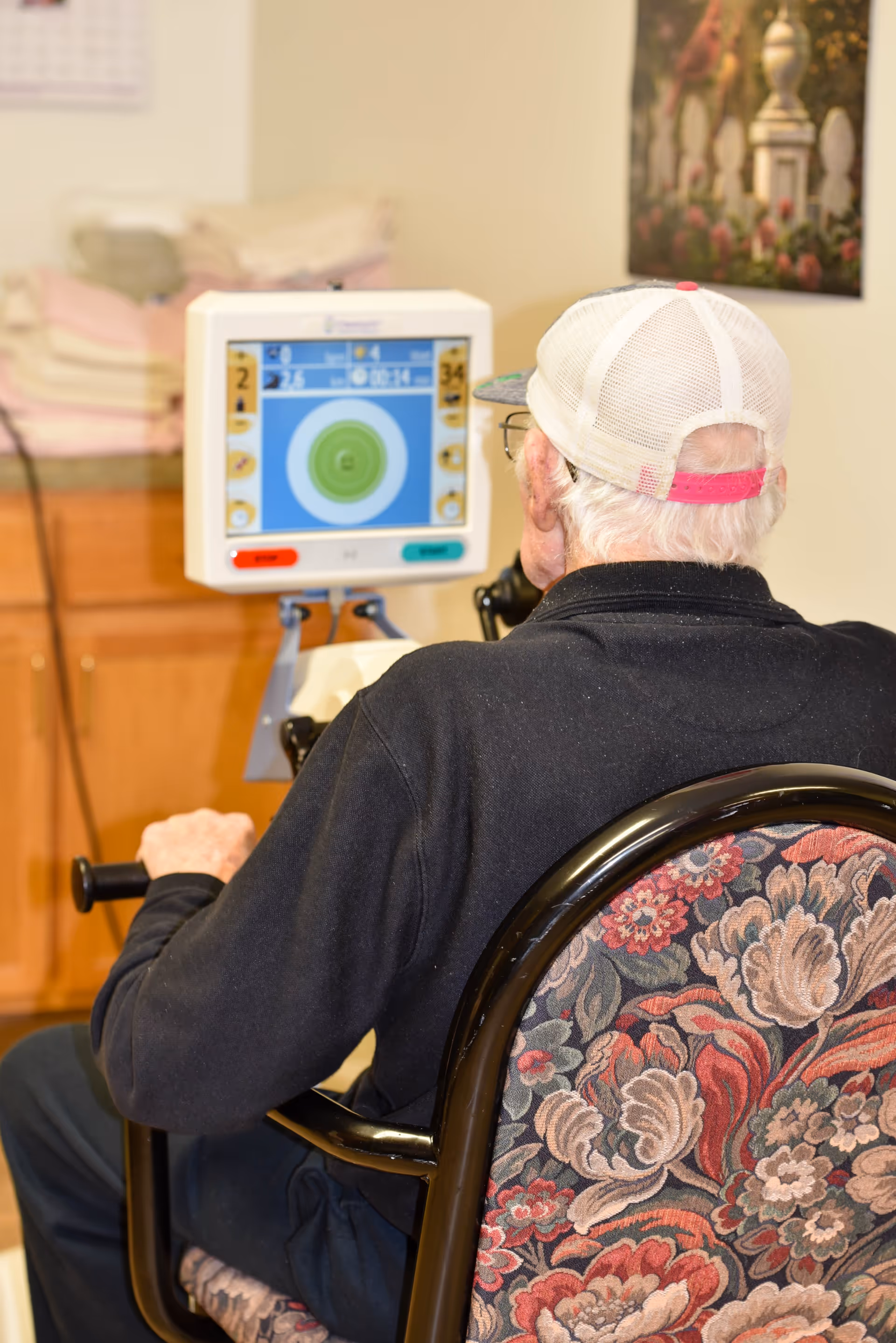 An elderly man wearing a white mesh cap with a pink strap is seated on a floral-patterned chair, using a piece of exercise equipment with a digital screen displaying a target and various data. The setting appears to be indoors with wooden cabinets and folded towels in the background.